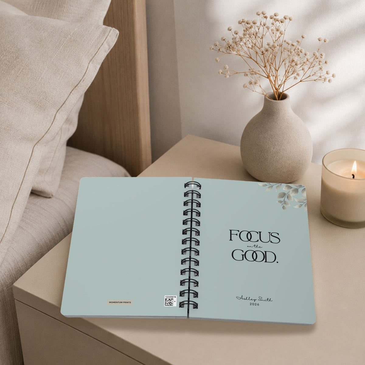 The opening spread of a personalized pale sky spiral notebook on a bedroom nightstand, showing the Momentum Prints tag and production code on the inside cover beside the title page with black lettering and a script name personalization, styled with a candle and dried baby's breath.