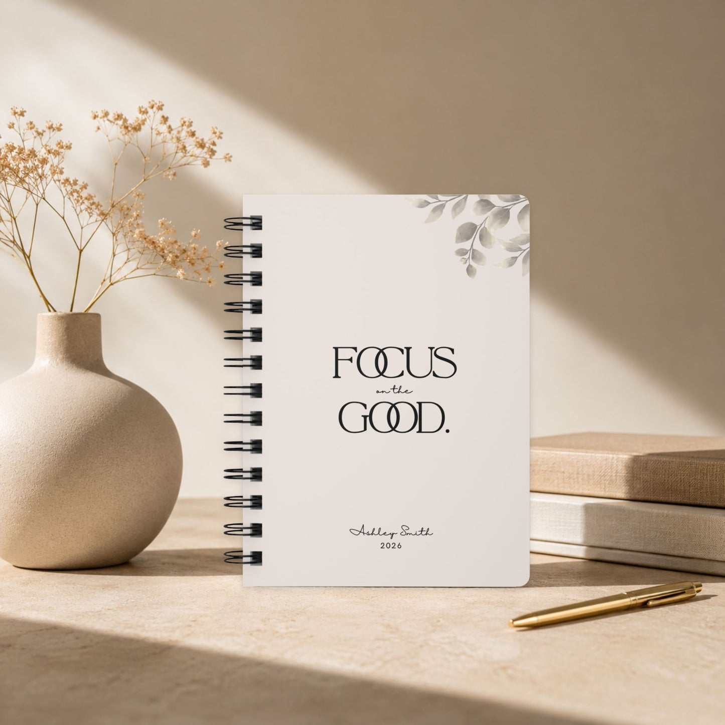 A personalized soft beige spiral notebook standing upright against a sunlit wall, framed by dried baby's breath in a stoneware vase, a stack of linen bound books, and a gold pen.