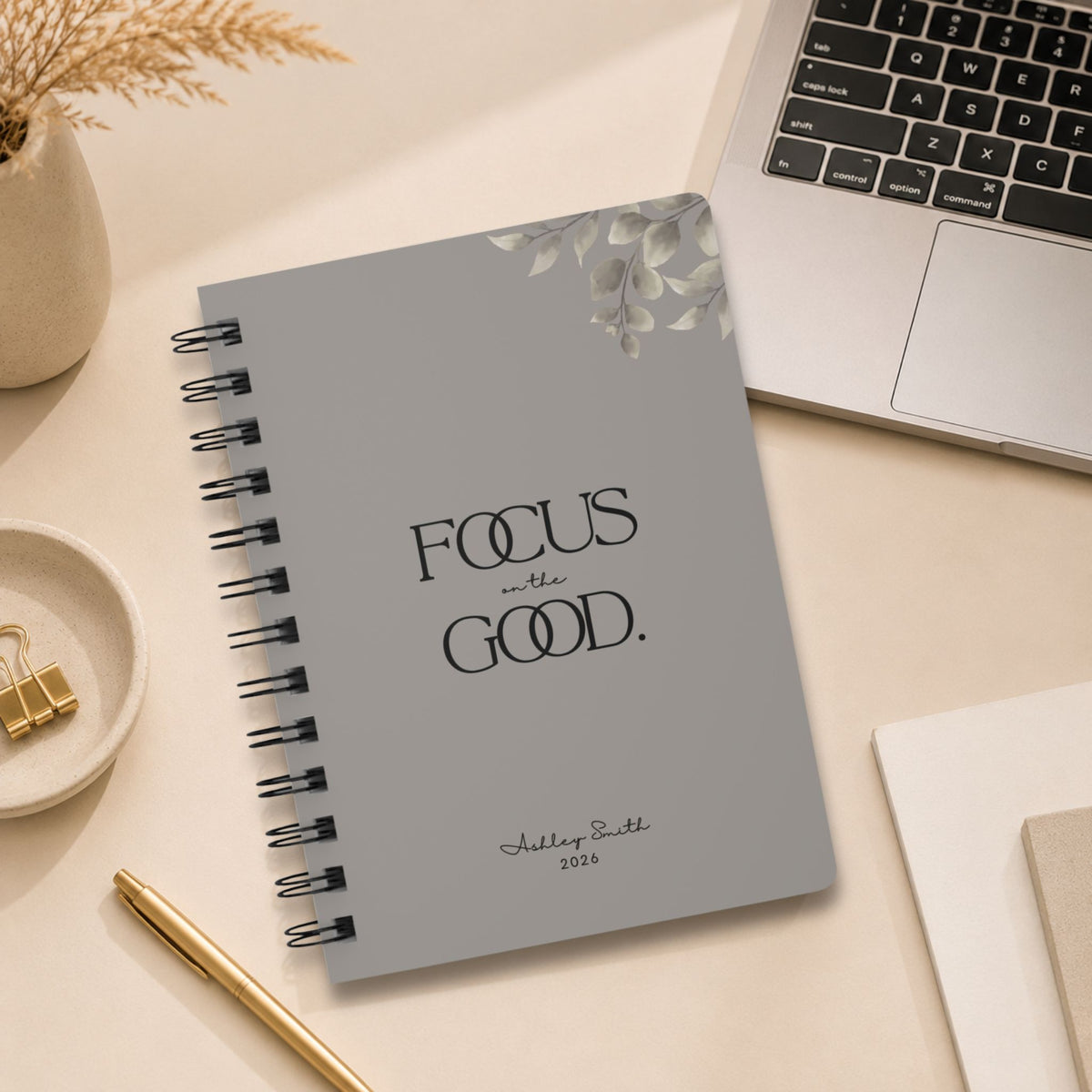 A personalized warm gray spiral notebook resting on a desk beside an open laptop, styled with dried pampas grass, a small dish of gold binder clips, and a gold pen.