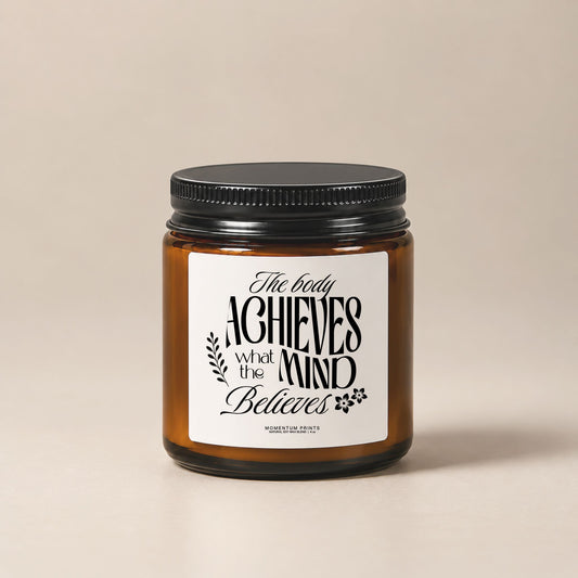 The Body Achieves What the Mind Believes 4 oz amber glass candle with black lid against a soft beige minimalist background.