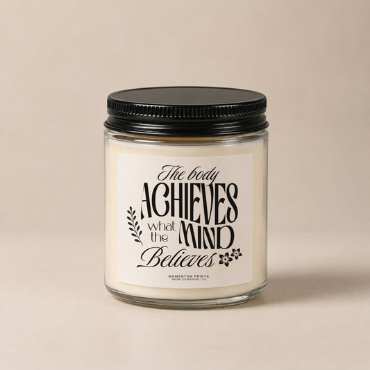 The Body Achieves What the Mind Believes 9 oz clear glass candle with black lid against a soft beige minimalist background.
