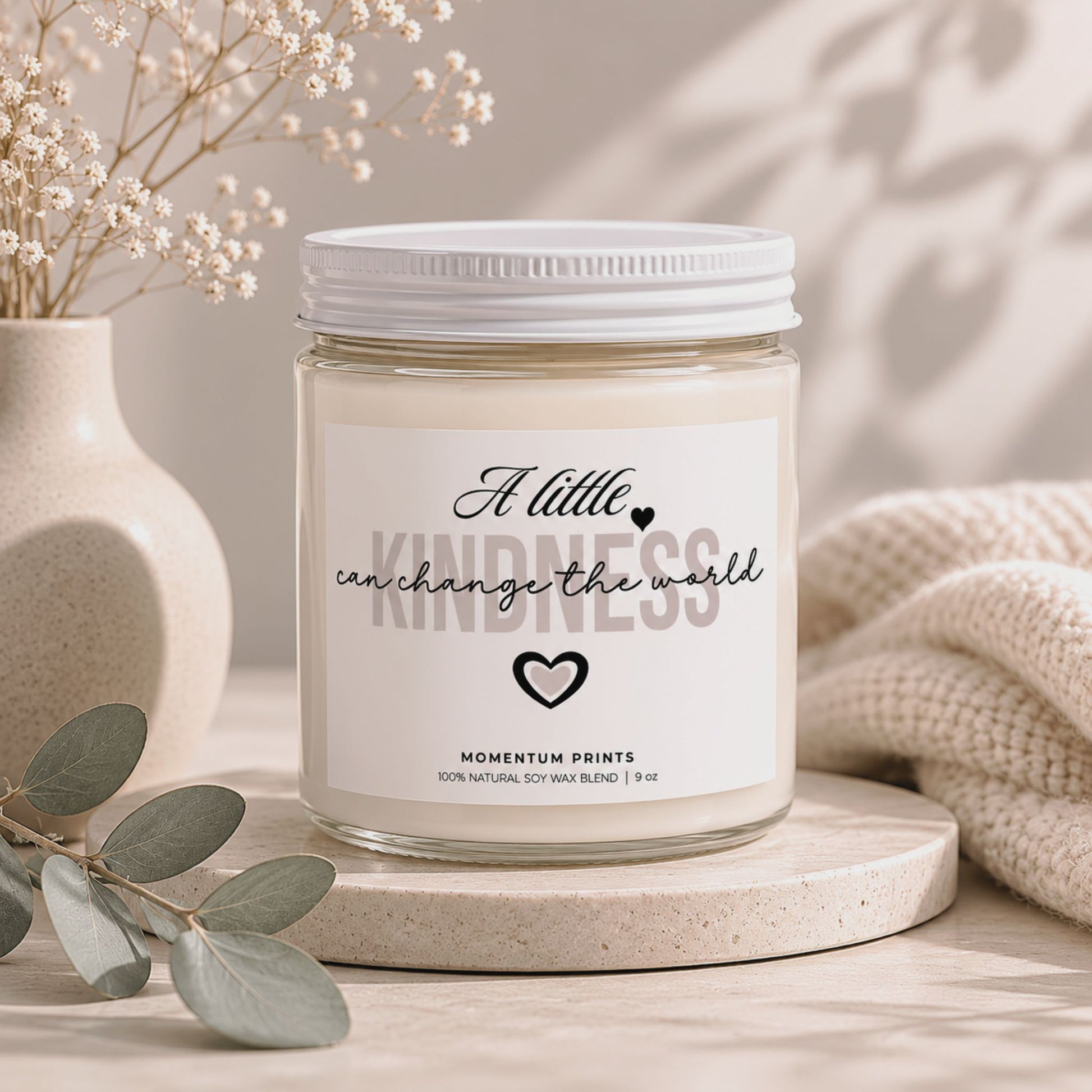A Little Kindness Can Change the World scented candle by Momentum Prints styled with eucalyptus leaves, soft natural light, and neutral beige decor.