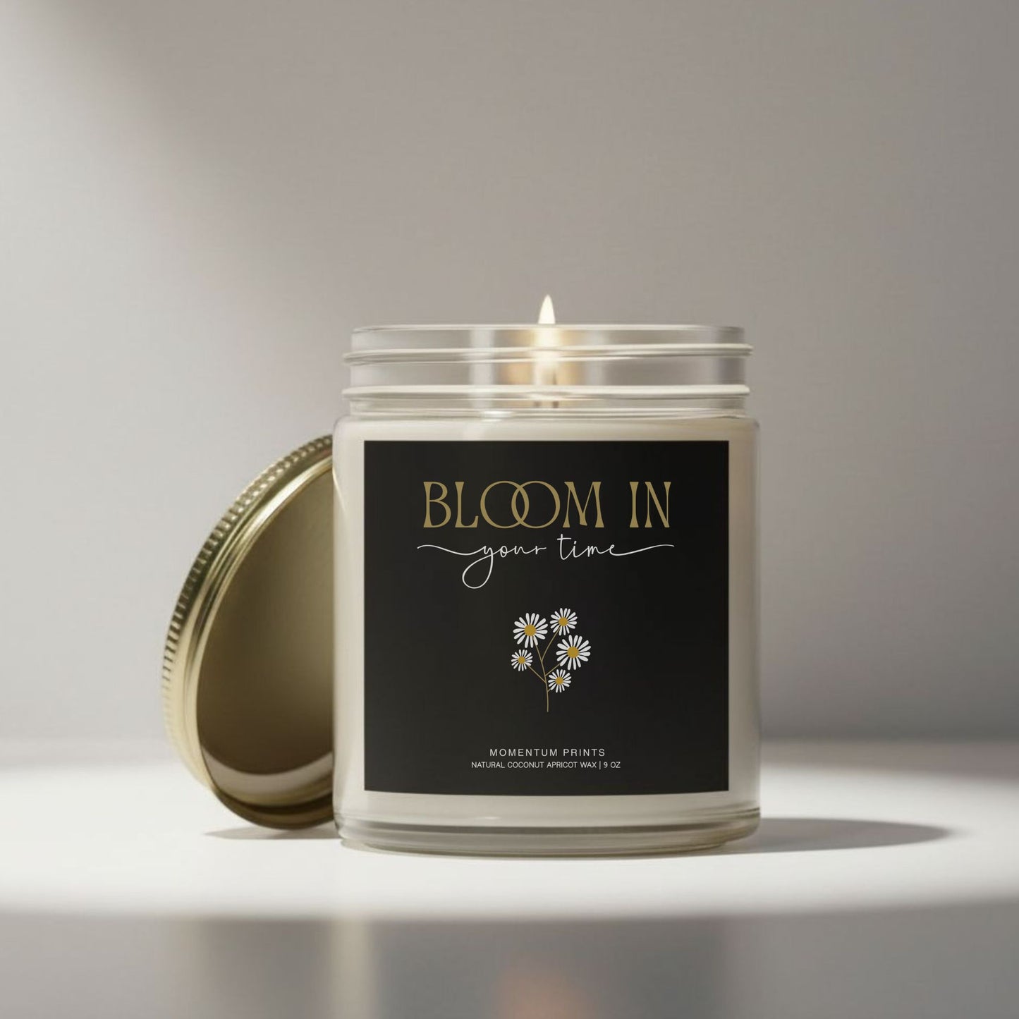 “Bloom in Your Time” black-label motivational candle on neutral minimalist background with soft natural light — daisy floral design by Momentum Prints.