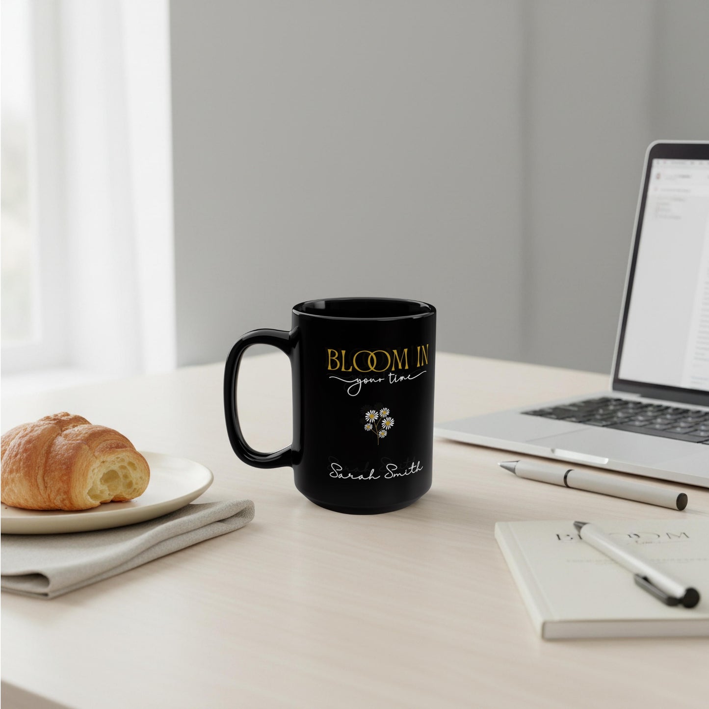 Personalized black ceramic mug with “Bloom in Your Time” design on a minimalist desk with laptop, croissant, and neutral stationery.