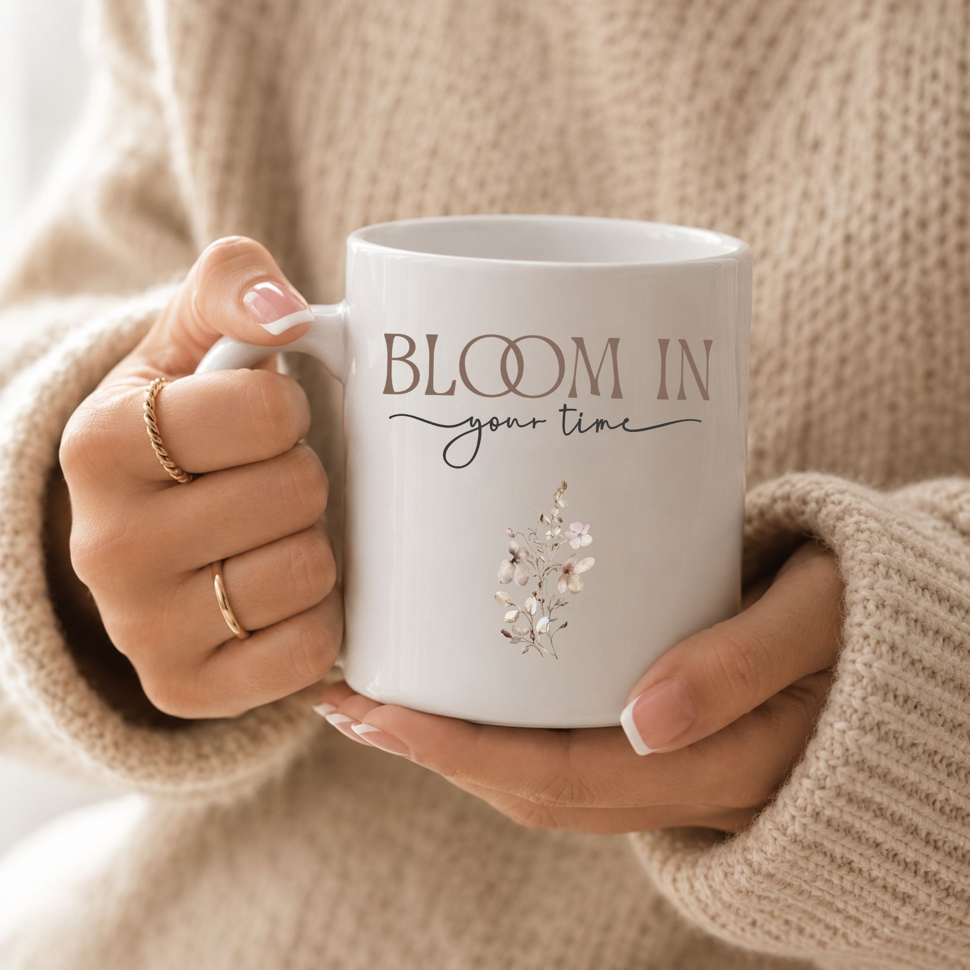 Woman holding a Bloom in Your Time ceramic mug while wearing a cozy beige knit sweater, creating a warm and inviting lifestyle scene.