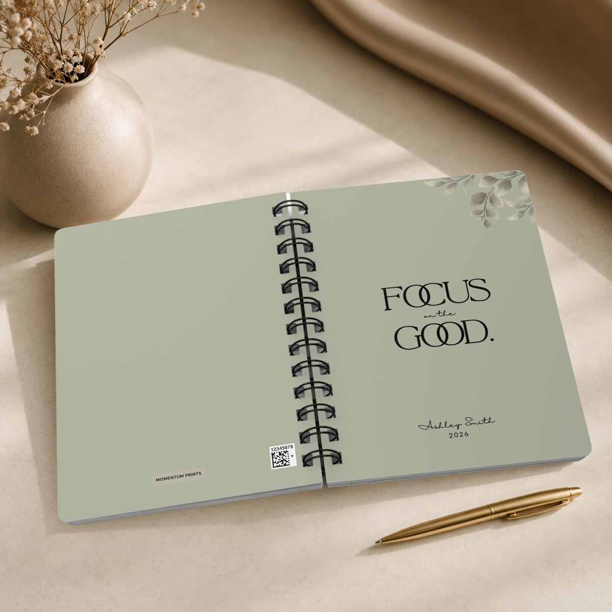 The opening spread of a personalized dried sage spiral notebook, showing the Momentum Prints tag and production code on the inside cover beside the title page with black lettering and a script name personalization. 
