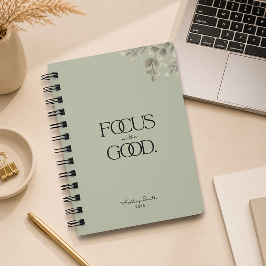 A personalized dried sage spiral notebook standing upright beside an open laptop, styled with dried pampas grass, gold binder clips, and a gold pen. Momentum Prints.