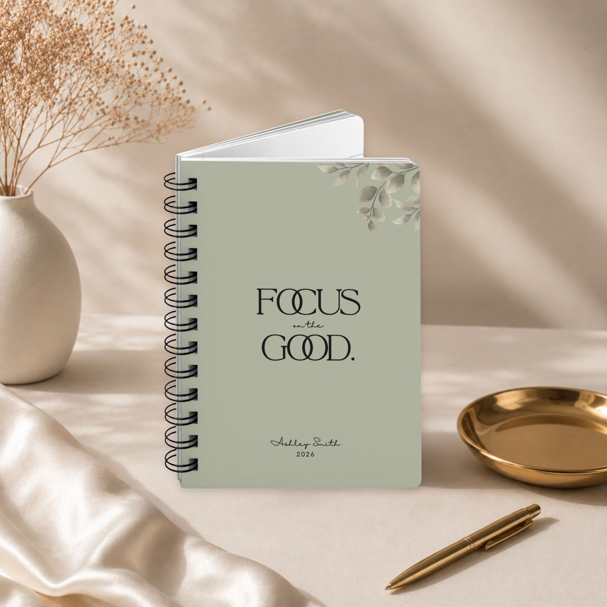 A personalized dried sage spiral notebook standing slightly open to reveal its page block, styled with dried baby's breath, a gold dish, and a gold pen.