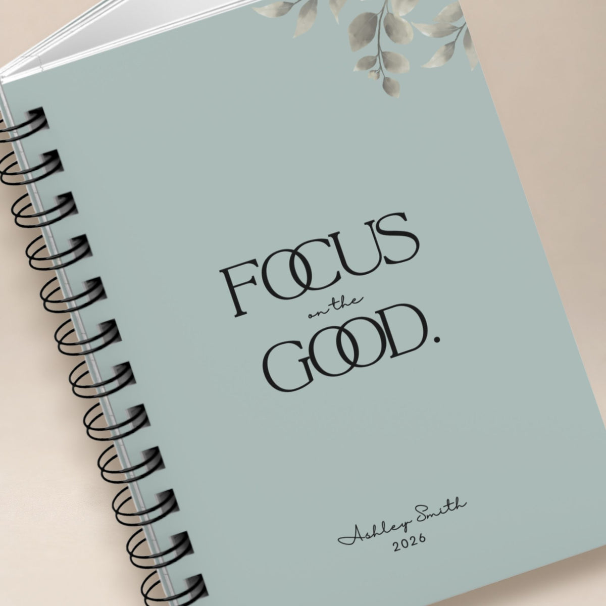 Close-up detail of a personalized pale sky spiral notebook cover, showing the black lettering, muted botanical illustration, and script name personalization. Momentum Prints.