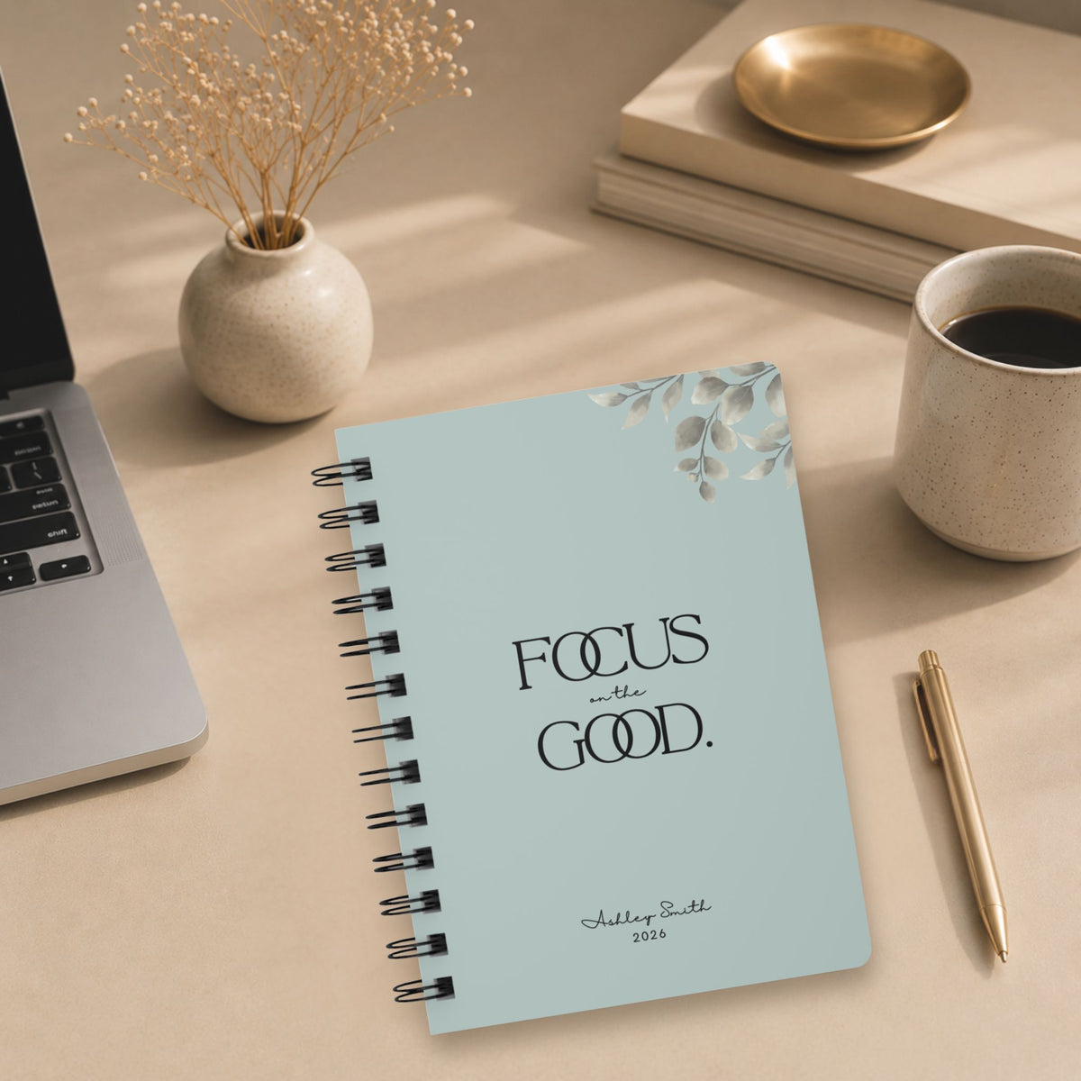 A personalized pale sky spiral notebook resting on a desk beside an open laptop, styled with dried baby's breath, a mug of coffee, stacked books, and a gold pen.