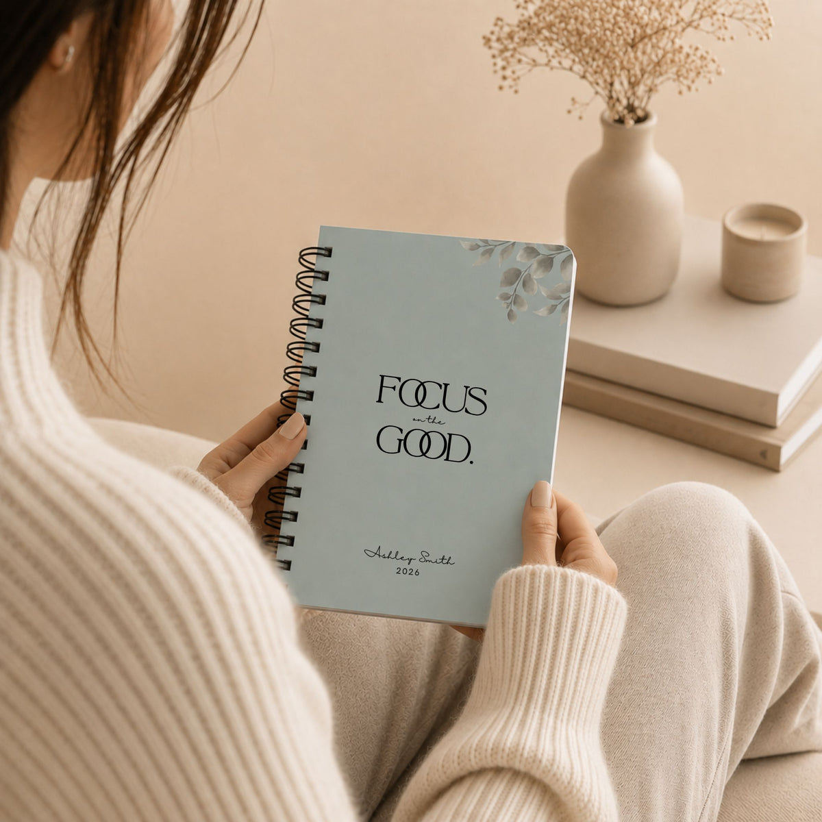 A woman in a cream sweater holding a personalized pale sky spiral notebook while sitting curled up on a couch, styled with dried baby's breath, a candle, and stacked books in the background.