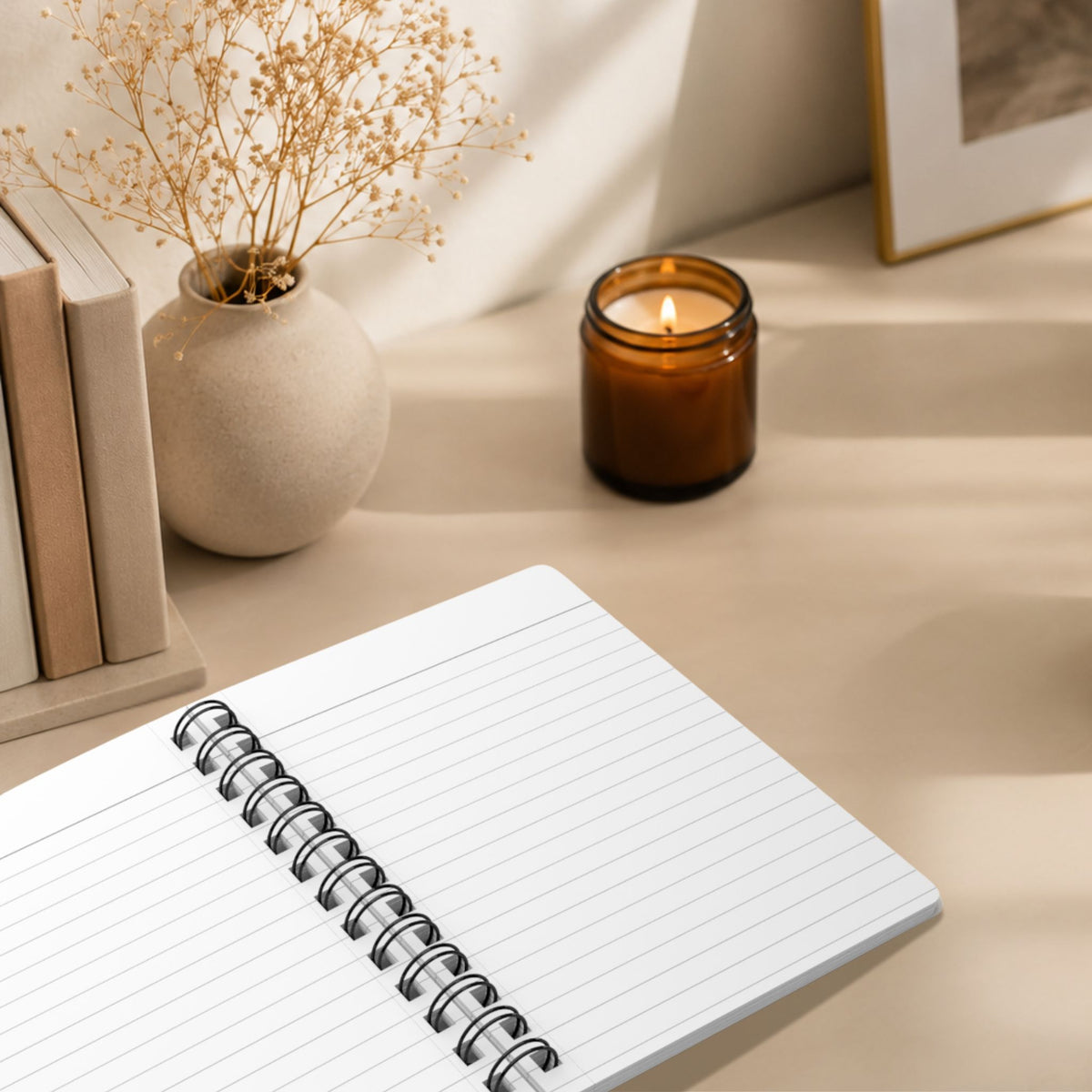 The blank lined pages of a personalized pale sky spiral notebook laid open on a desk, styled with a lit amber glass candle, stacked books, dried baby's breath, and a framed print in the background.