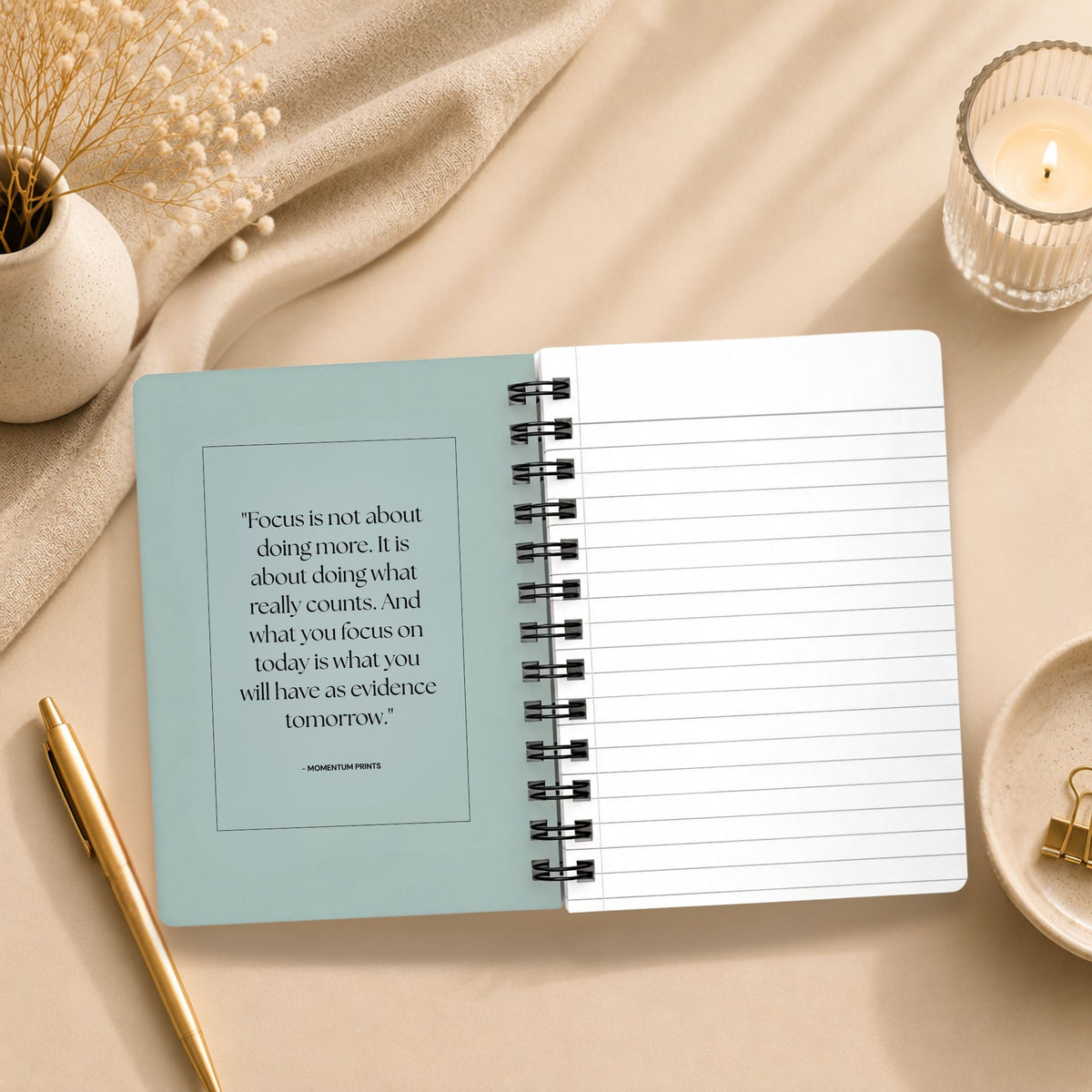 The open interior of a personalized pale sky spiral notebook, showing a printed reflection on focus and intention beside blank lined pages, styled with a lit candle, dried baby's breath, and a gold pen. Momentum Prints.