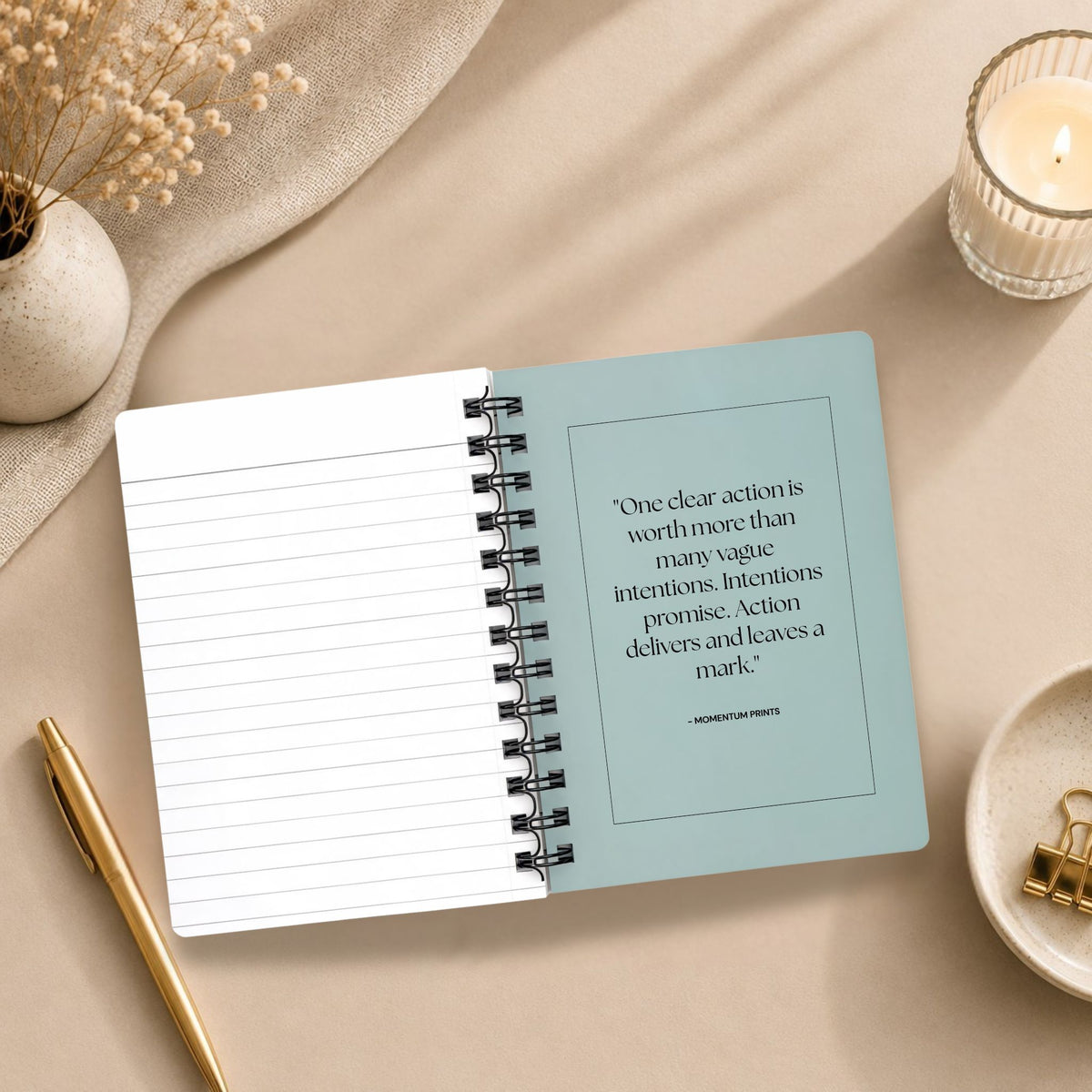 The open interior of a personalized pale sky spiral notebook, showing a blank lined page beside a printed message about taking clear action, styled with a lit candle, dried baby's breath, and gold binder clips. Momentum Prints.