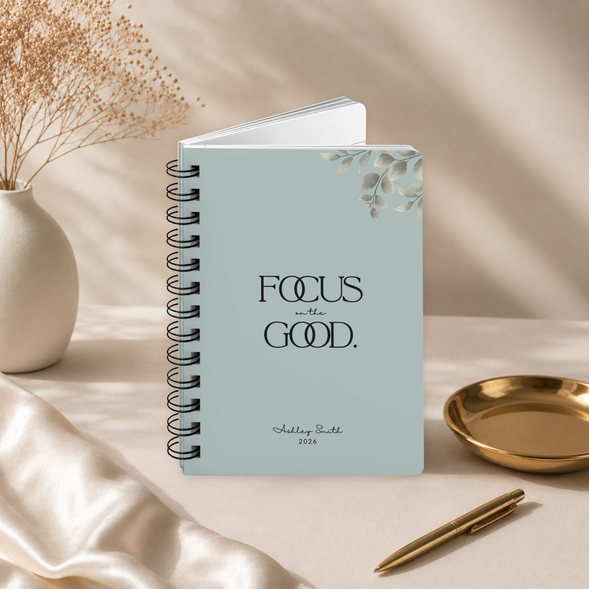 A personalized pale sky spiral notebook standing slightly open to reveal its page block, styled with dried baby's breath, a gold dish, and a gold pen.