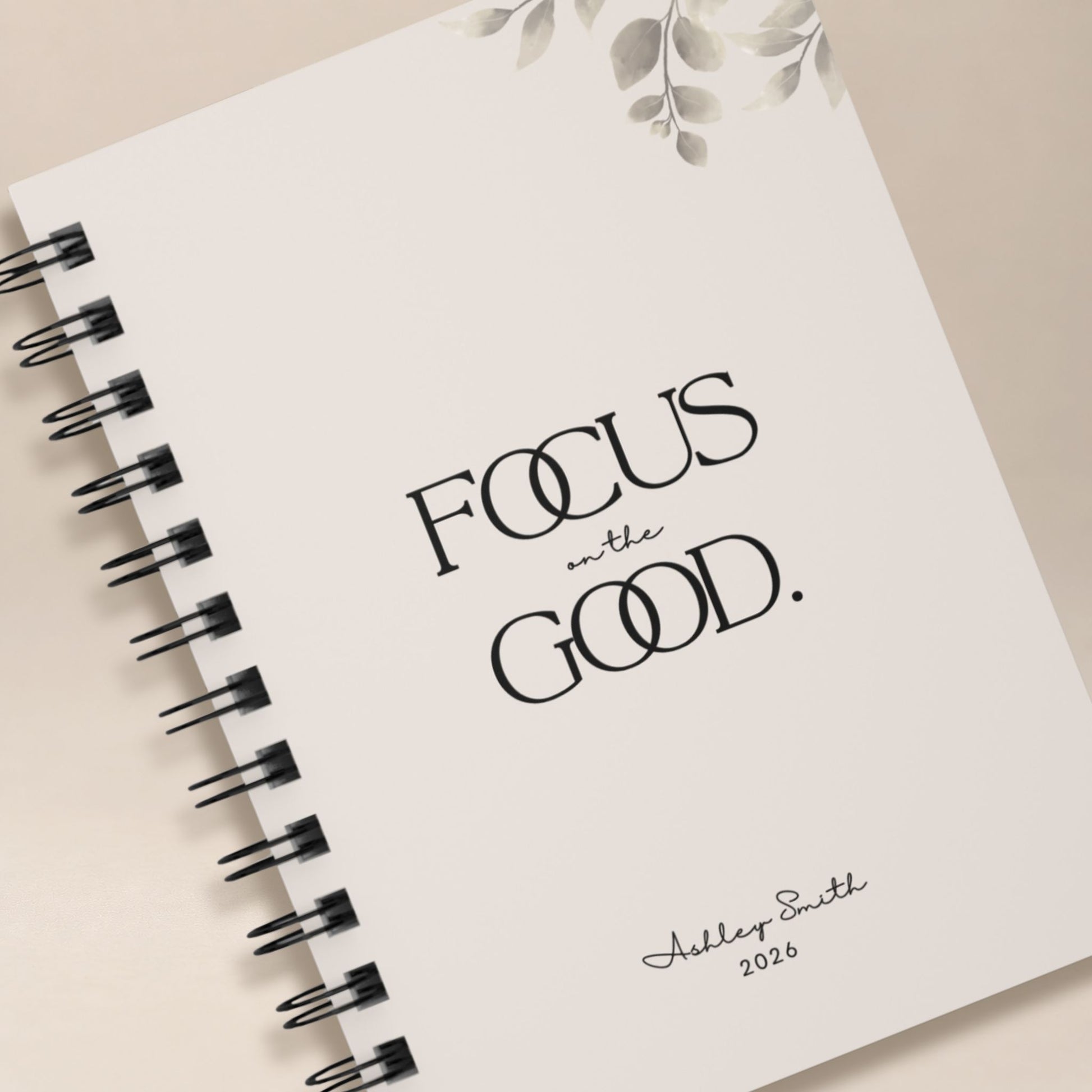 Close up detail of a personalized soft beige spiral notebook cover, showing the black lettering, muted botanical illustration, and script name personalization. Momentum Prints.