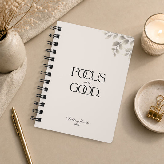A personalized soft beige spiral notebook styled in an overhead flat lay with dried botanical stems, a lit candle, gold binder clips on a small ceramic dish, and a gold pen resting on warm neutral linen.