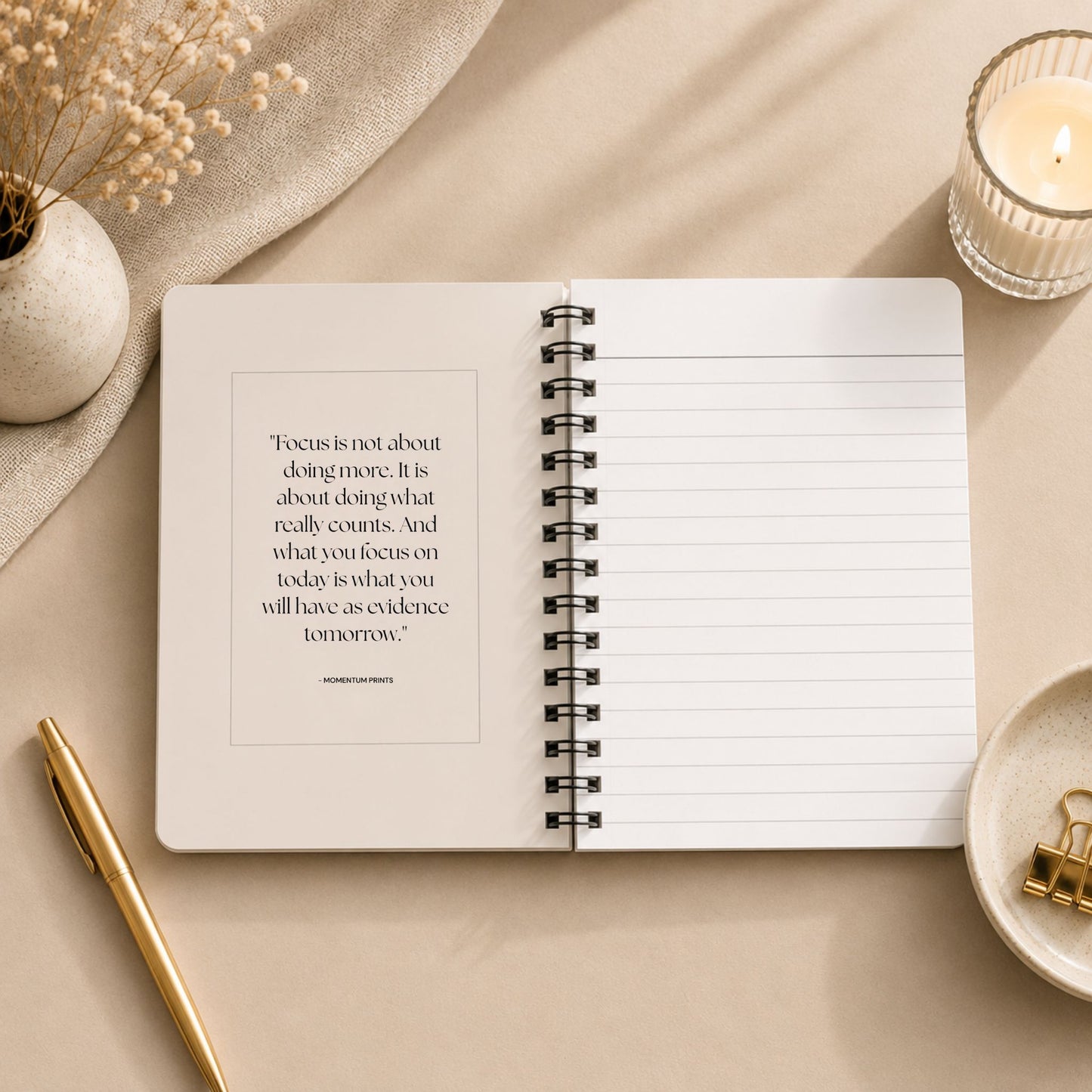 The open interior of a personalized soft beige spiral notebook, showing a printed reflection on focus and intention beside blank lined pages, styled with a lit glass candle, dried baby's breath on draped linen, and a gold pen.