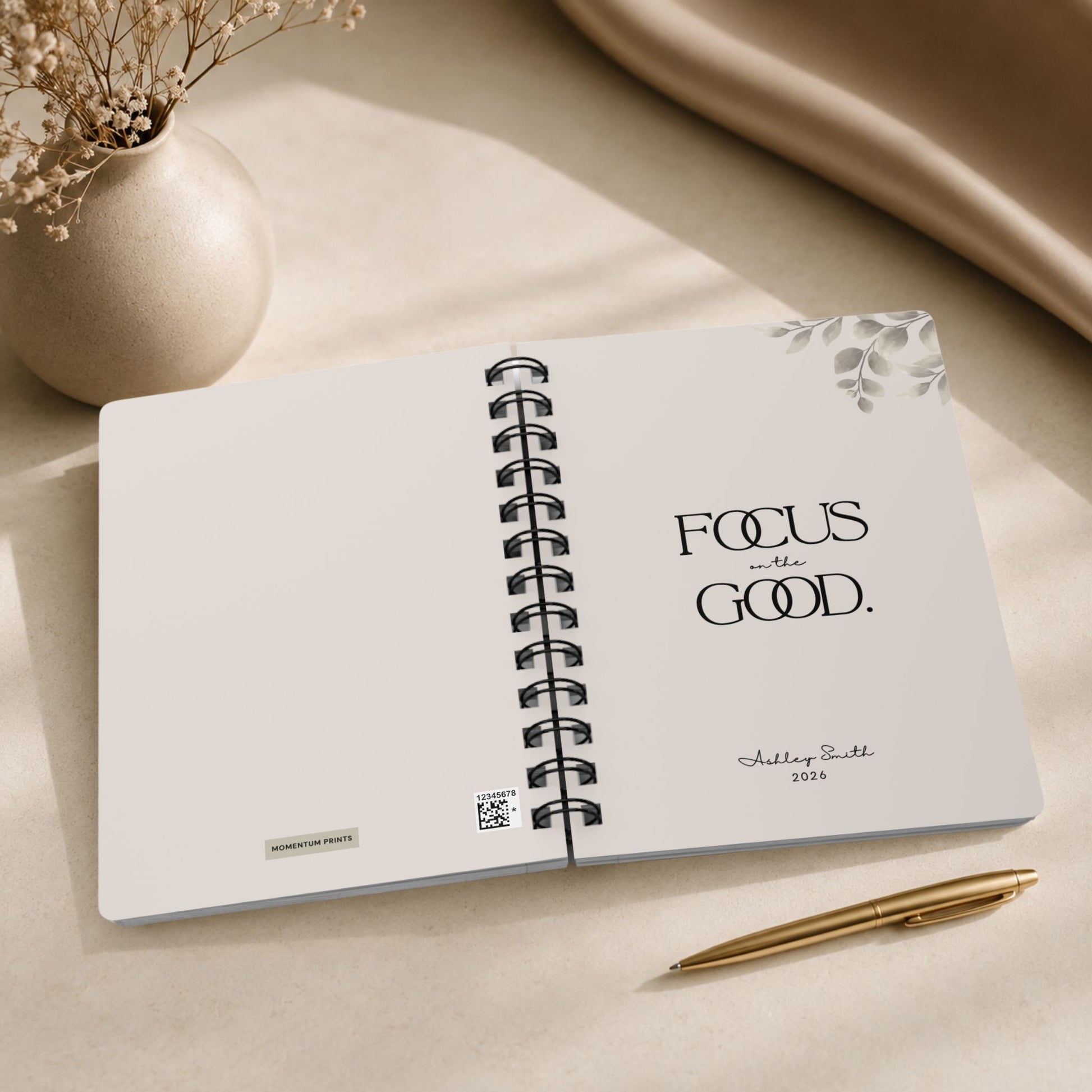 The opening spread of a personalized soft beige spiral notebook, showing the Momentum Prints tag and production code on the inside cover beside the title page with black lettering and a script name personalization.