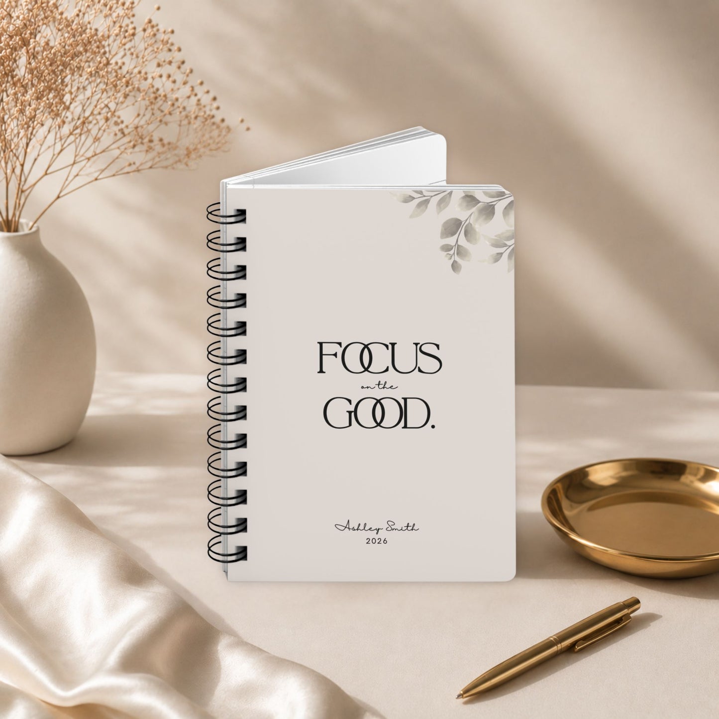 A personalized soft beige spiral notebook standing slightly open to reveal its page block, styled with a gold pen, a small gold dish, and draped silk fabric in warm light.