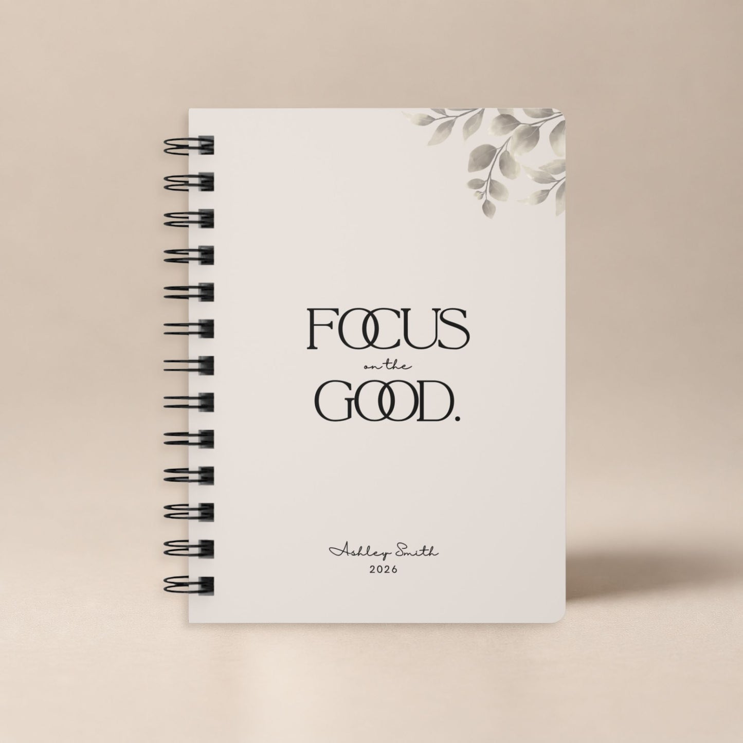 A personalized soft beige spiral notebook standing upright against a plain warm neutral backdrop, shown as a clean product hero shot. Momentum Prints.
