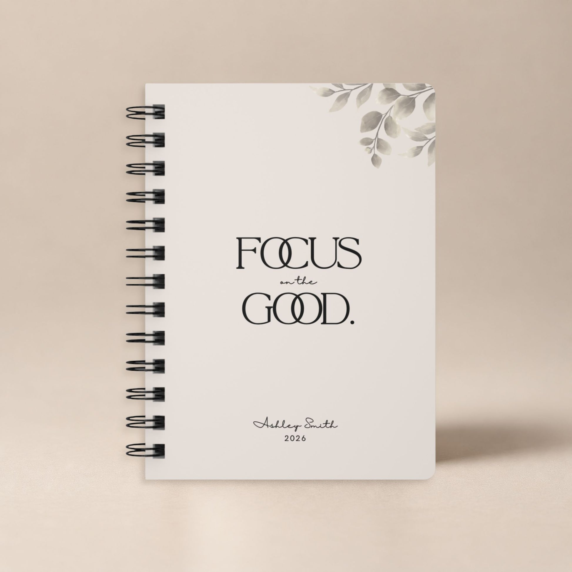 A personalized soft beige spiral notebook standing upright against a plain warm neutral backdrop, shown as a clean product hero shot. Momentum Prints.