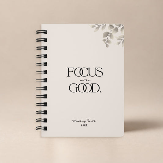 A personalized soft beige spiral notebook standing upright against a plain warm neutral backdrop, shown as a clean product hero shot. Momentum Prints.