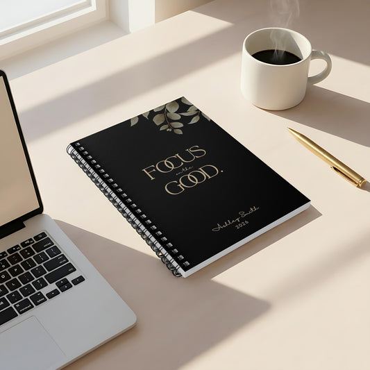 Momentum Prints motivational spiral notebook (6x8) in Midnight Black featuring the “Focus on the Good” cover on a minimalist desk with coffee and soft window light.