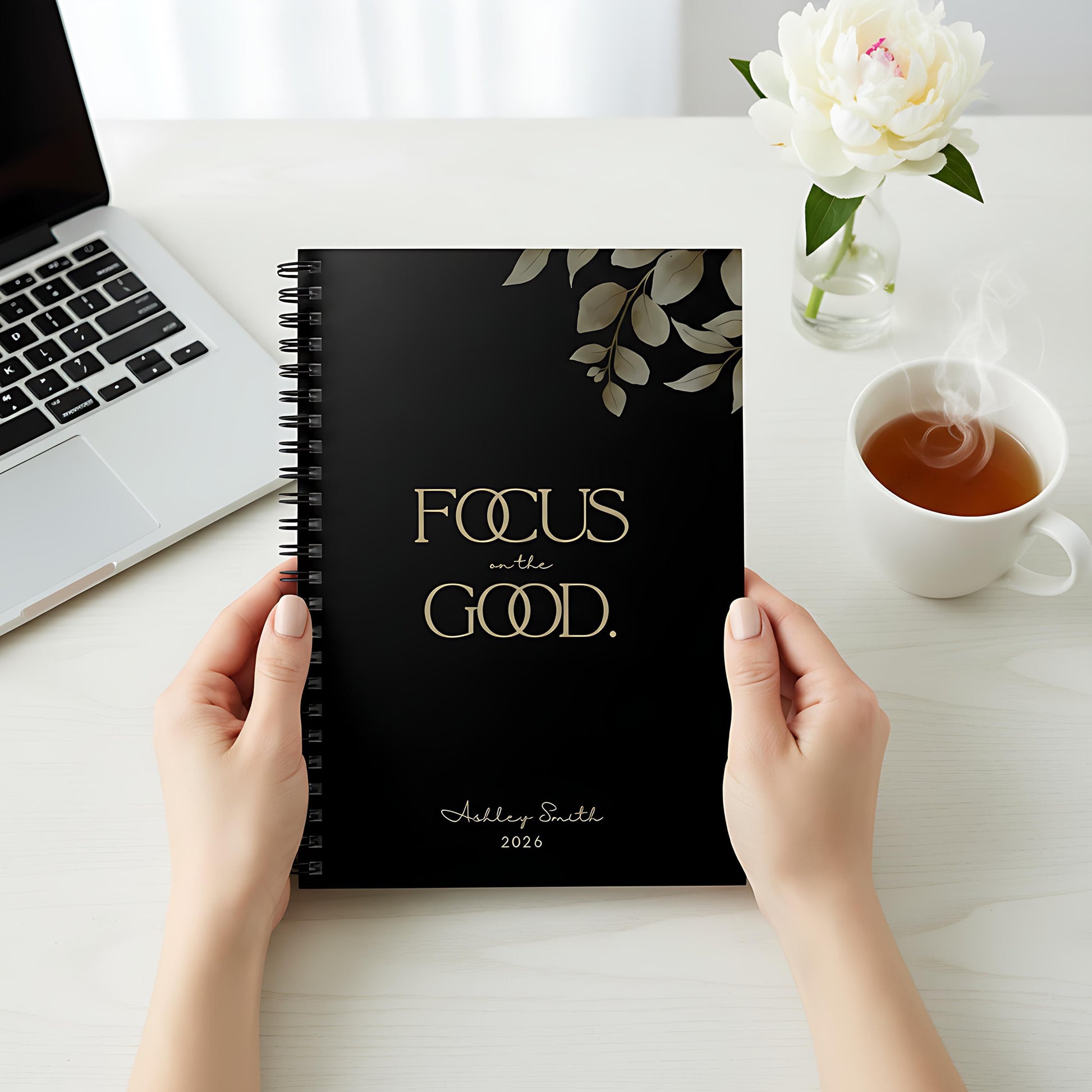 Hands holding a Momentum Prints spiral notebook (6x8) in Midnight Black with the “Focus on the Good” cover on a bright desk.