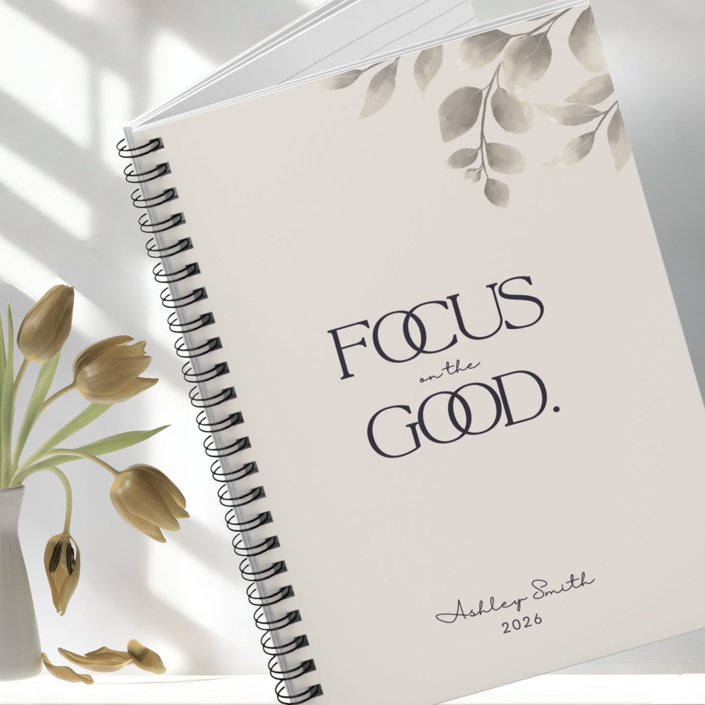 Angled close-up of soft beige spiral notebook with minimalist botanical details and elegant typography