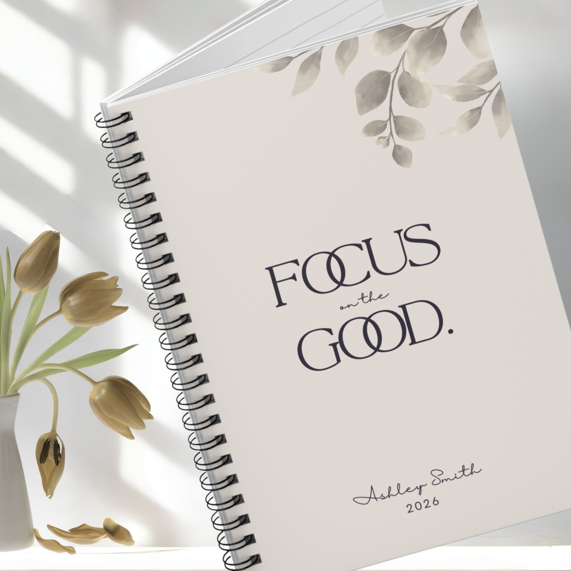 Angled close-up of soft beige spiral notebook with minimalist botanical details and elegant typography
