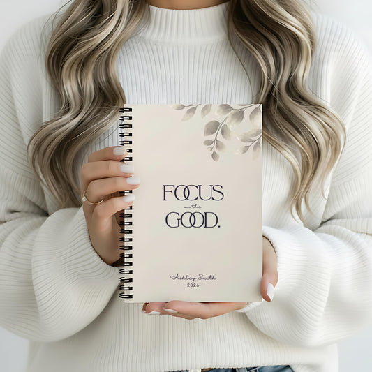 Woman holding a soft beige spiral notebook with the phrase “Focus on the Good” in a minimal cozy setting