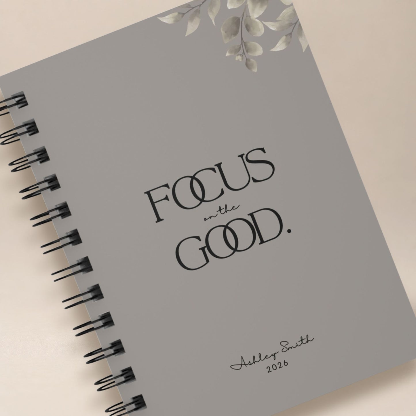 Close-up detail of a personalized warm gray spiral notebook cover, showing the black lettering, muted botanical illustration, and script name personalization.