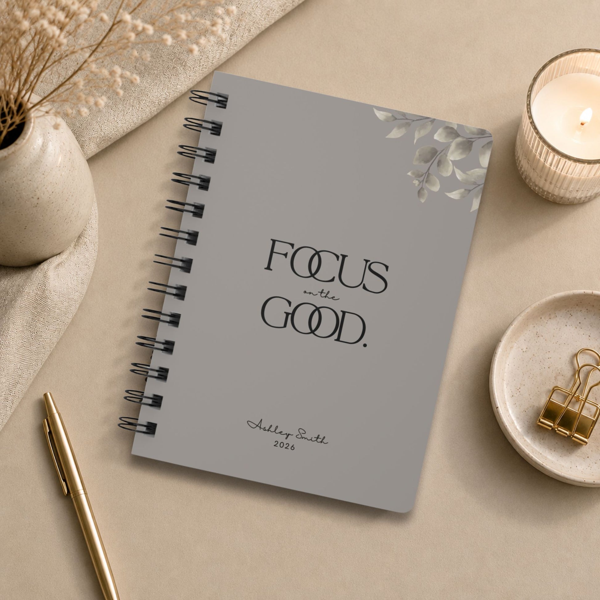 A personalized warm gray spiral notebook in a flat lay with draped linen, dried baby's breath, a lit glass candle, a small dish of gold binder clips, and a gold pen.