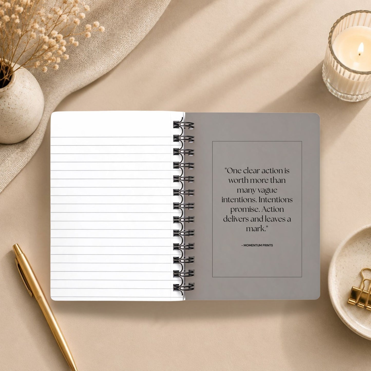 The open interior of a personalized warm gray spiral notebook, showing a blank lined page beside a printed message about taking clear action, styled with a lit glass candle, dried baby's breath on draped linen, and a gold pen. Momentum Prints.