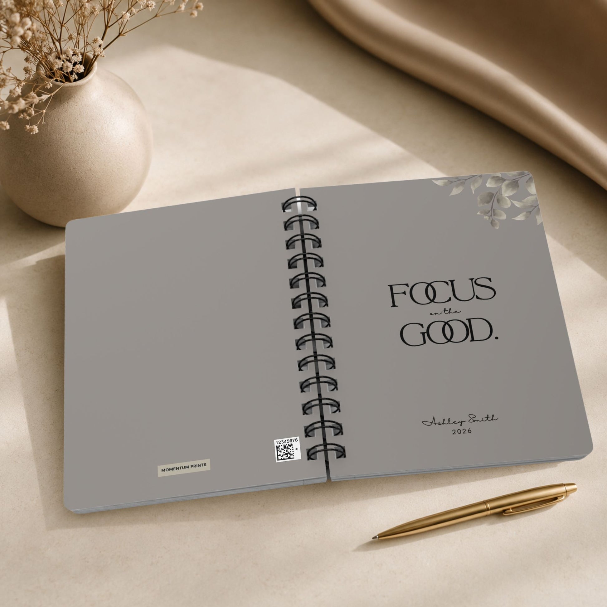 The opening spread of a personalized warm gray spiral notebook, showing the Momentum Prints tag and production code on the inside cover beside the title page with black lettering and a script name personalization.