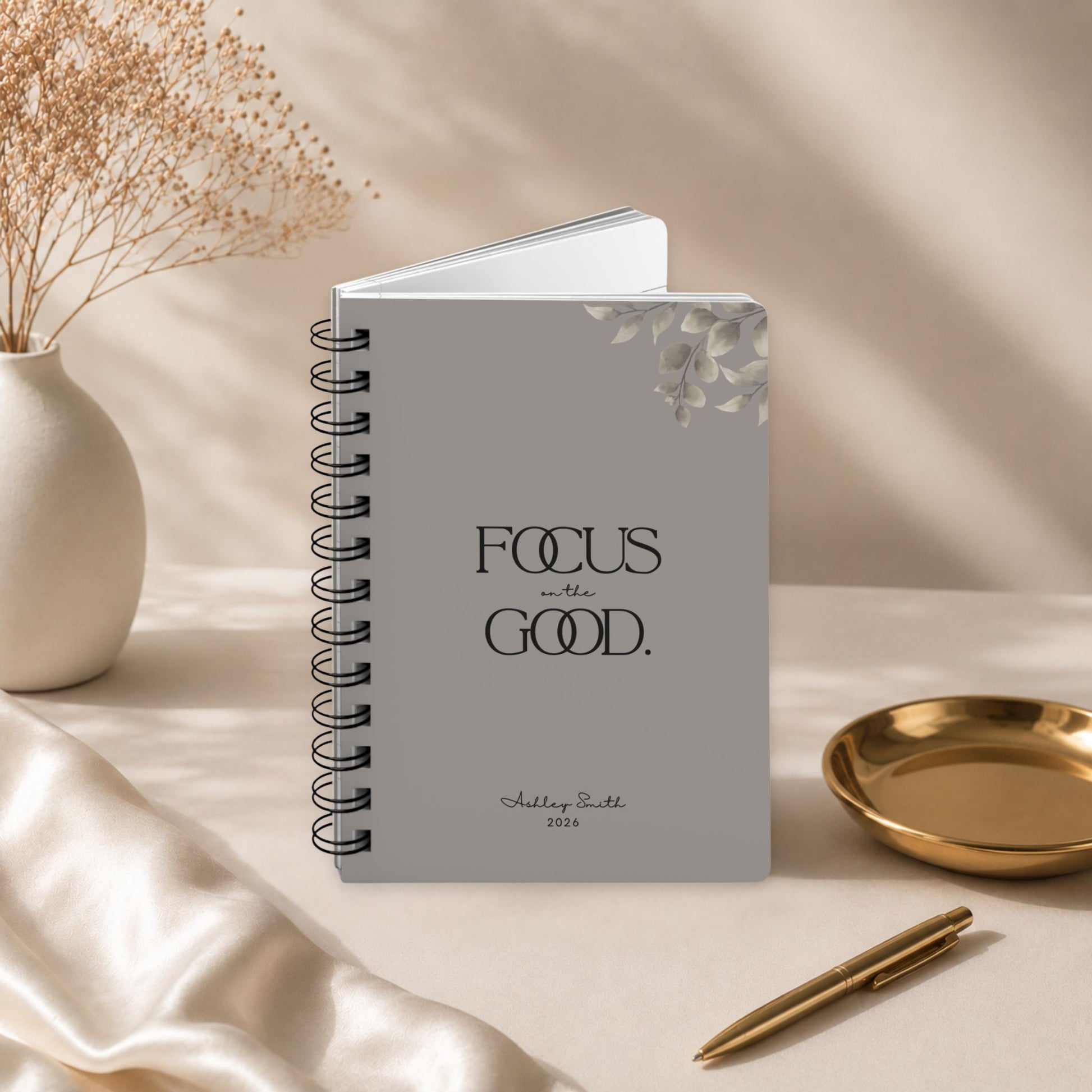 A personalized warm gray spiral notebook standing slightly open to reveal its page block, styled with draped silk fabric, dried baby's breath, and a gold dish. Momentum Prints.