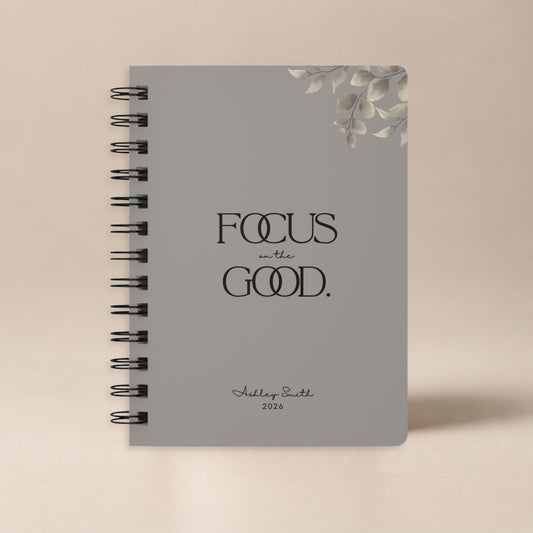 A personalized warm gray spiral notebook standing upright against a plain warm neutral backdrop, shown as a clean product hero shot.