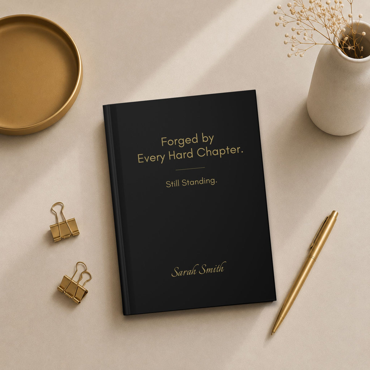 Minimal flat lay featuring a black personalized hardcover journal with gold binder clips, matte gold tray, ceramic vase, and gold pen.