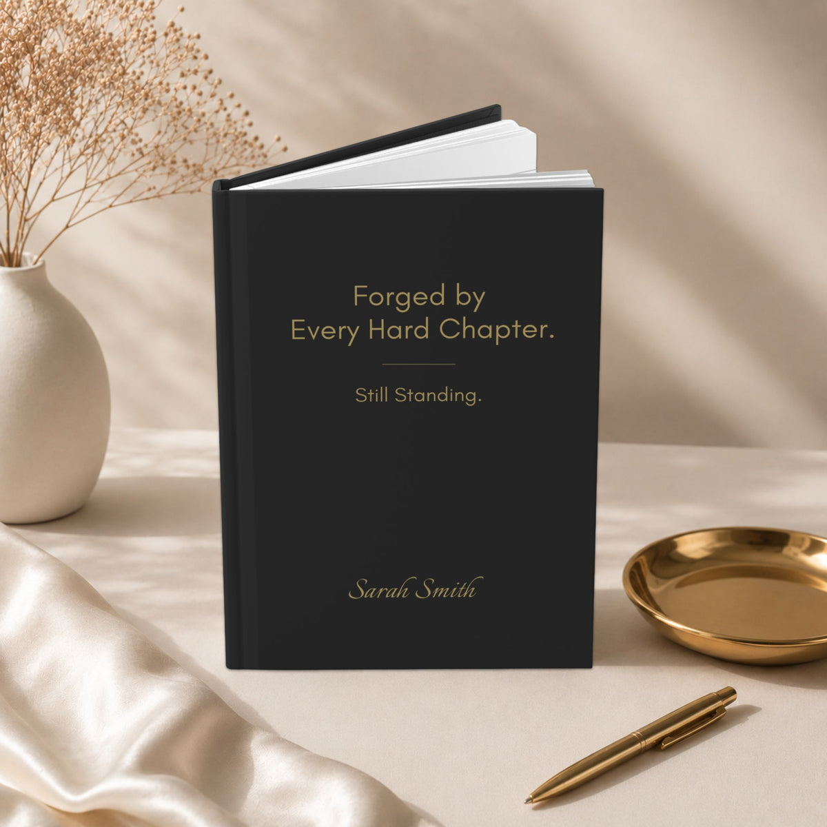 Black hardcover journal standing upright with the quote "Forged by Every Hard Chapter. Still Standing." styled in a minimalist workspace with a matte gold tray and neutral decor.