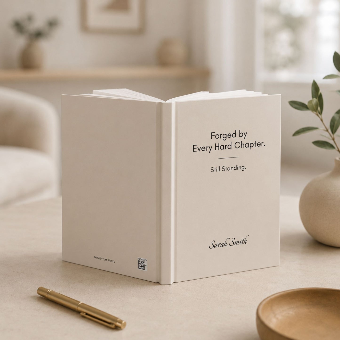An open ivory hardcover journal standing upright, showing its spine with the Momentum Prints wordmark, in a softly blurred living room setting.