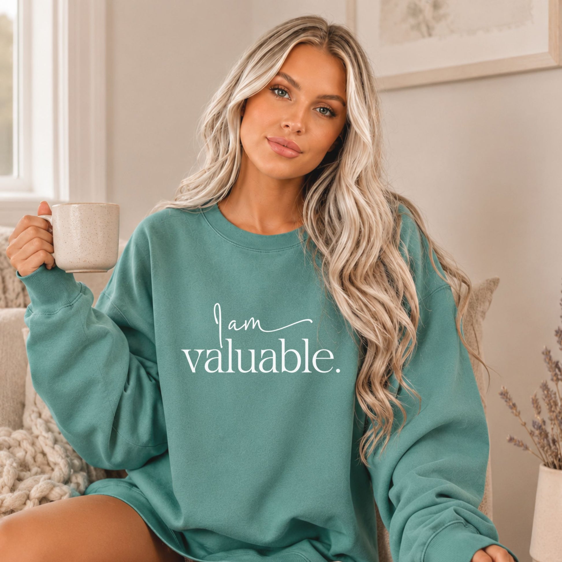 Woman wearing an oversized Light Green Comfort Colors sweatshirt with motivational affirmation design, holding a coffee mug in a cozy minimalist home setting. Relaxed lifestyle apparel mockup with neutral decor and warm natural light by Momentum Prints.