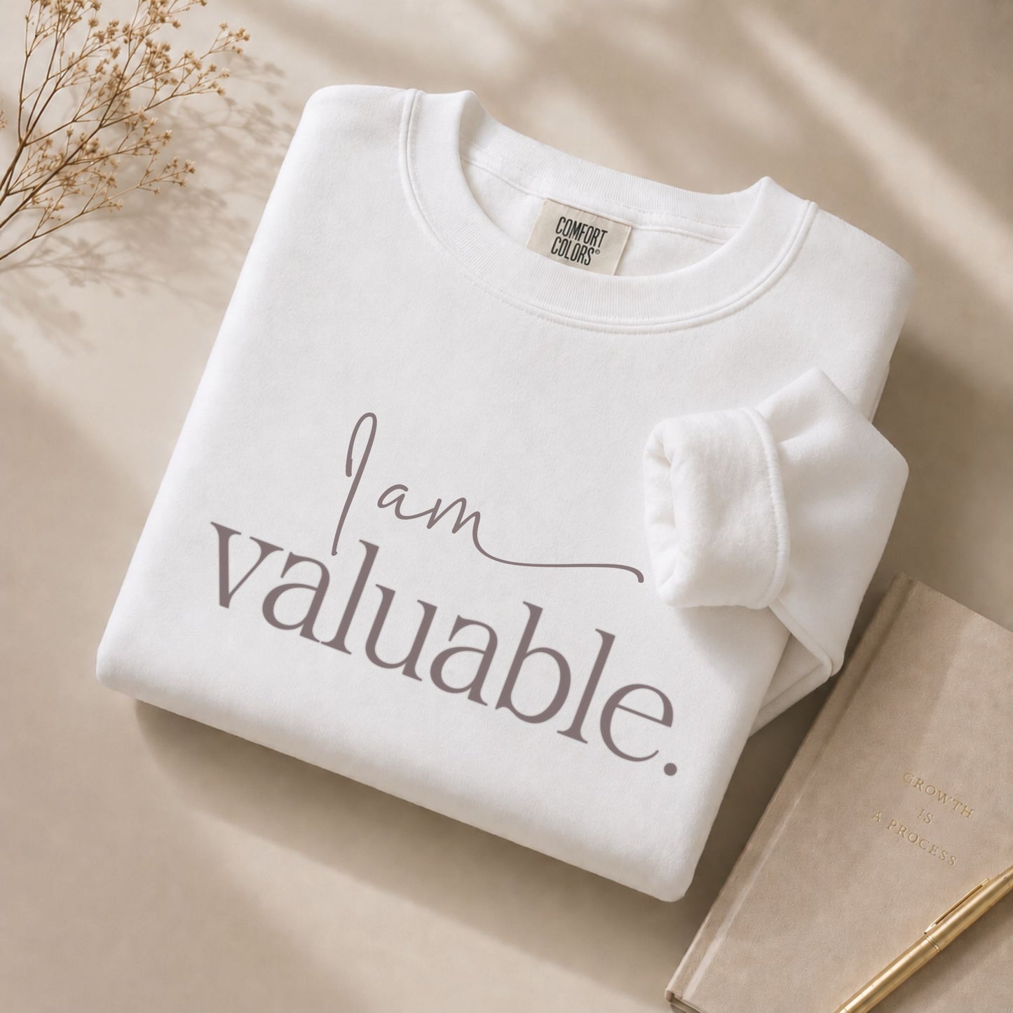 Close-up flat lay of the I Am Valuable white sweatshirt with visible Comfort Colors label, styled on a beige background with a journal and gold pen in a premium minimalist setting.