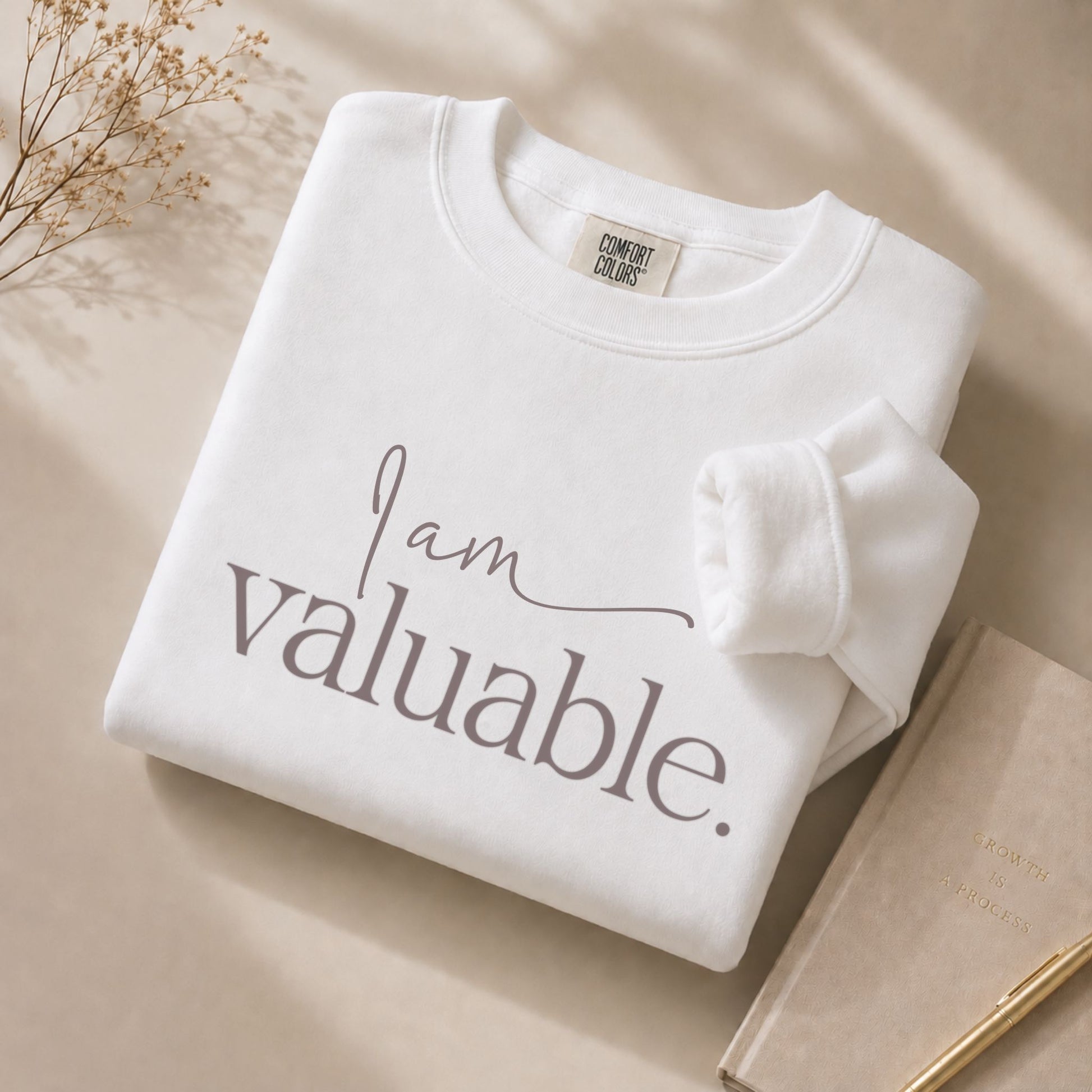 Close-up flat lay of the I Am Valuable white sweatshirt with visible Comfort Colors label, styled on a beige background with a journal and gold pen in a premium minimalist setting.