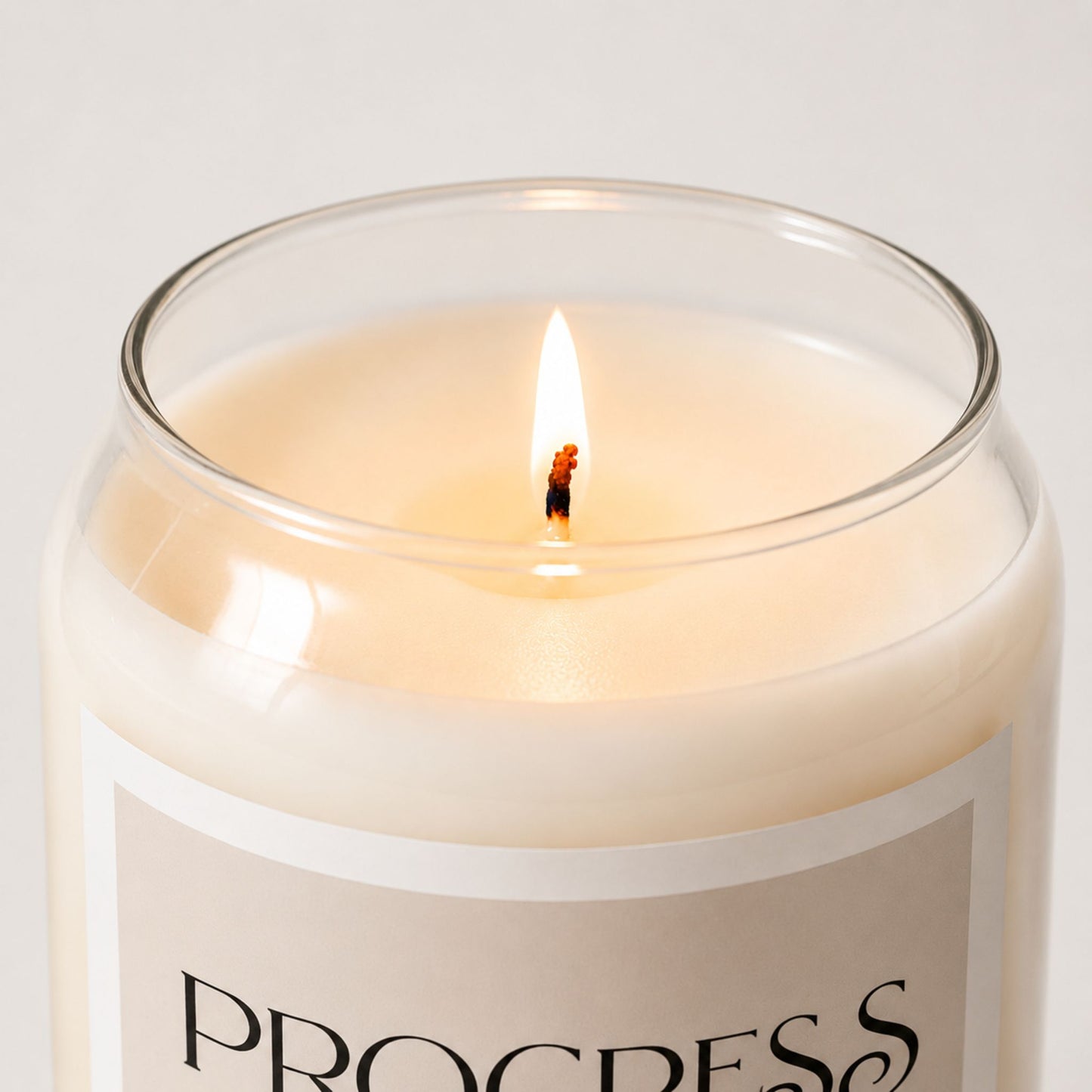 Close-up of a lit minimalist soy candle in a clear glass jar with creamy wax and a soft beige label with white border, styled in a clean neutral background with warm natural lighting.