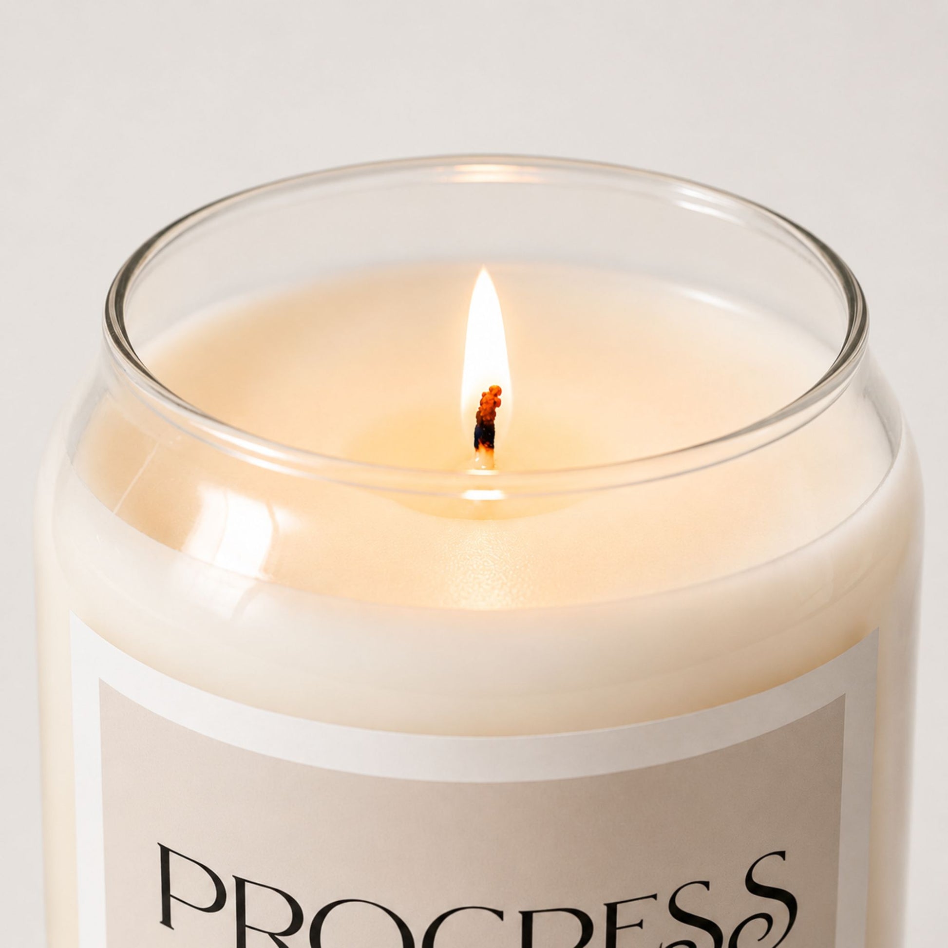 Close-up of a lit minimalist soy candle in a clear glass jar with creamy wax and a soft beige label with white border, styled in a clean neutral background with warm natural lighting.