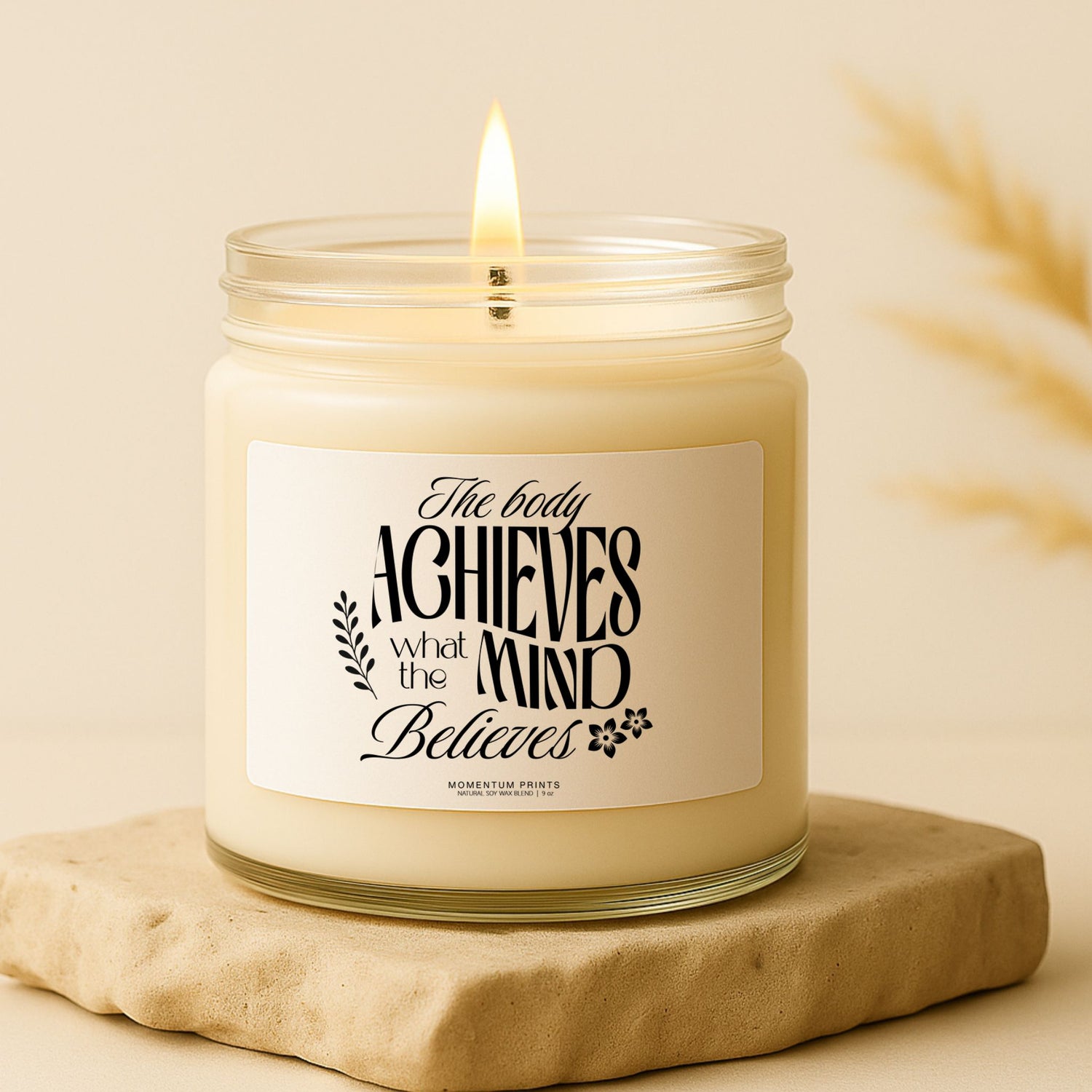 Motivational scented candle in clear jar with "The Body Achieves" label on beige stone base, Momentum Prints.