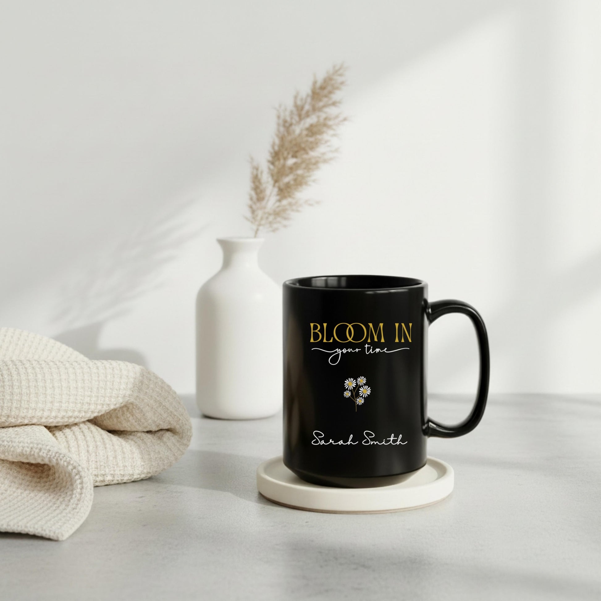Personalized black ceramic mug with “Bloom in Your Time” design in gold and white lettering, styled on a neutral tray with a white vase and soft knit blanket in a cozy minimalist setting.