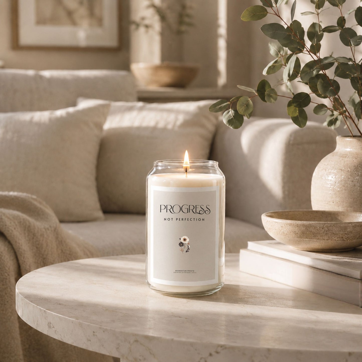 Progress Not Perfection 13.75 oz scented soy candle in clear glass jar with blank minimalist label, displayed on round marble coffee table in warm neutral living room with eucalyptus vase, ceramic bowl, and soft natural sunlight. Luxury lifestyle candle mockup by Momentum Prints.