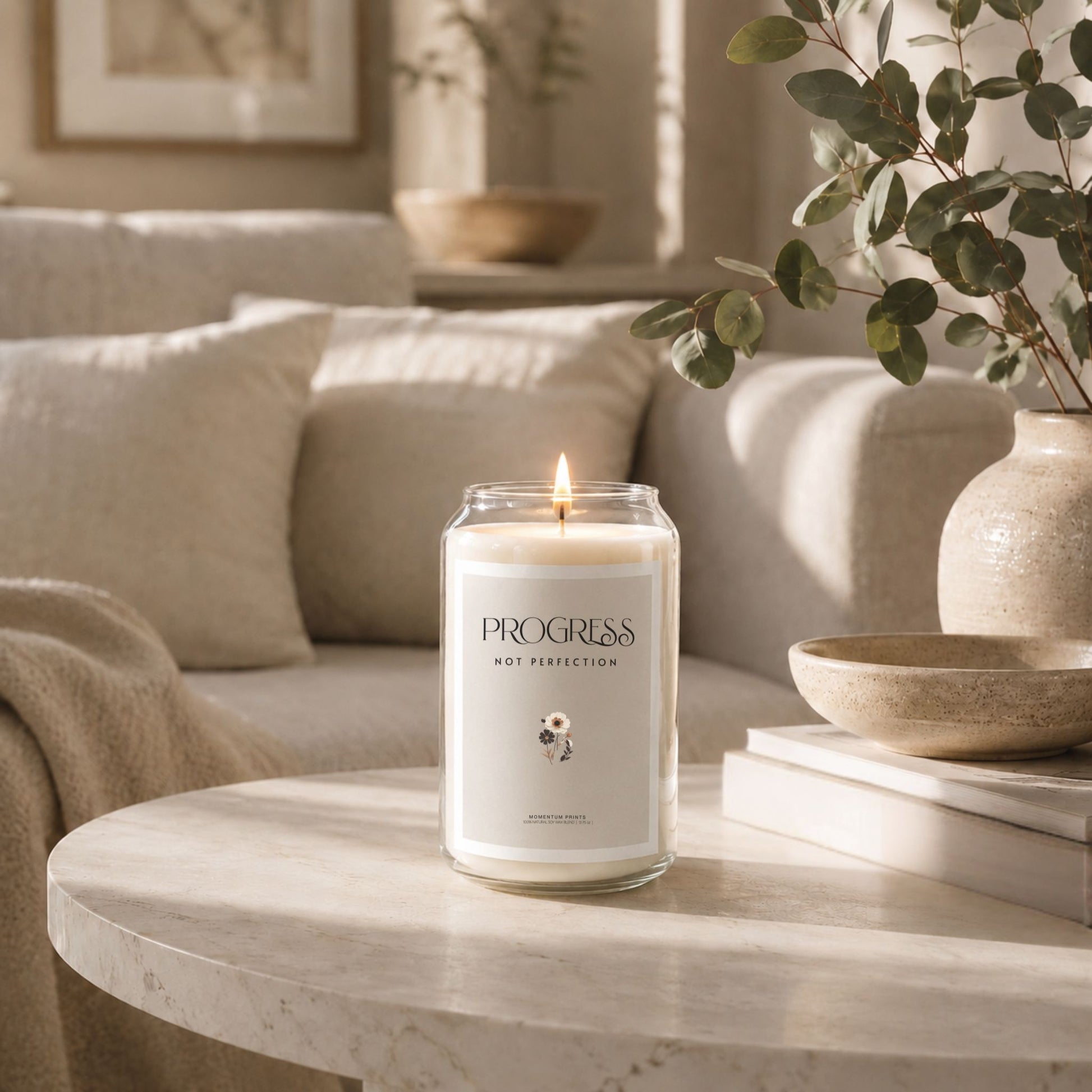 Progress Not Perfection 13.75 oz scented soy candle in clear glass jar with blank minimalist label, displayed on round marble coffee table in warm neutral living room with eucalyptus vase, ceramic bowl, and soft natural sunlight. Luxury lifestyle candle mockup by Momentum Prints.